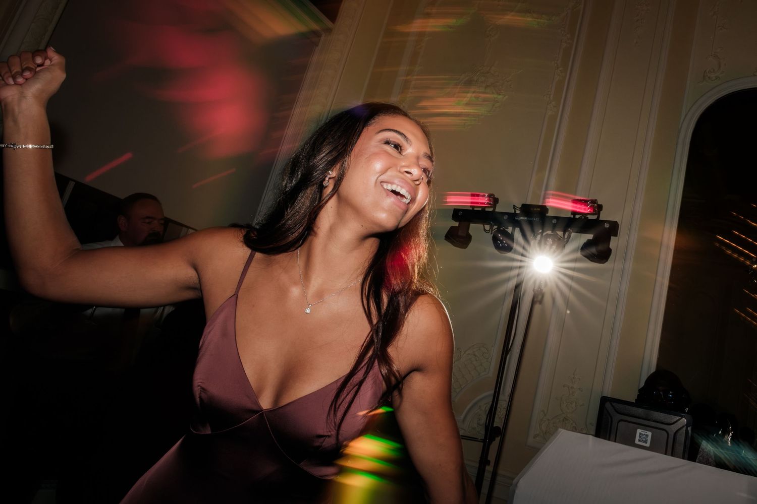 Smiling woman with long dark hair dances joyfully at an evening event, wearing a mauve dress with stage lights glowing behind her.