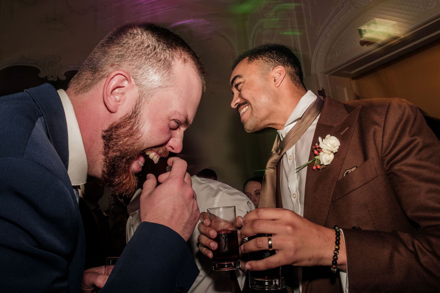 Two groomsmen laugh and drink whiskey at a wedding reception, one in a navy suit and the other in a brown jacket with a white boutonniere.