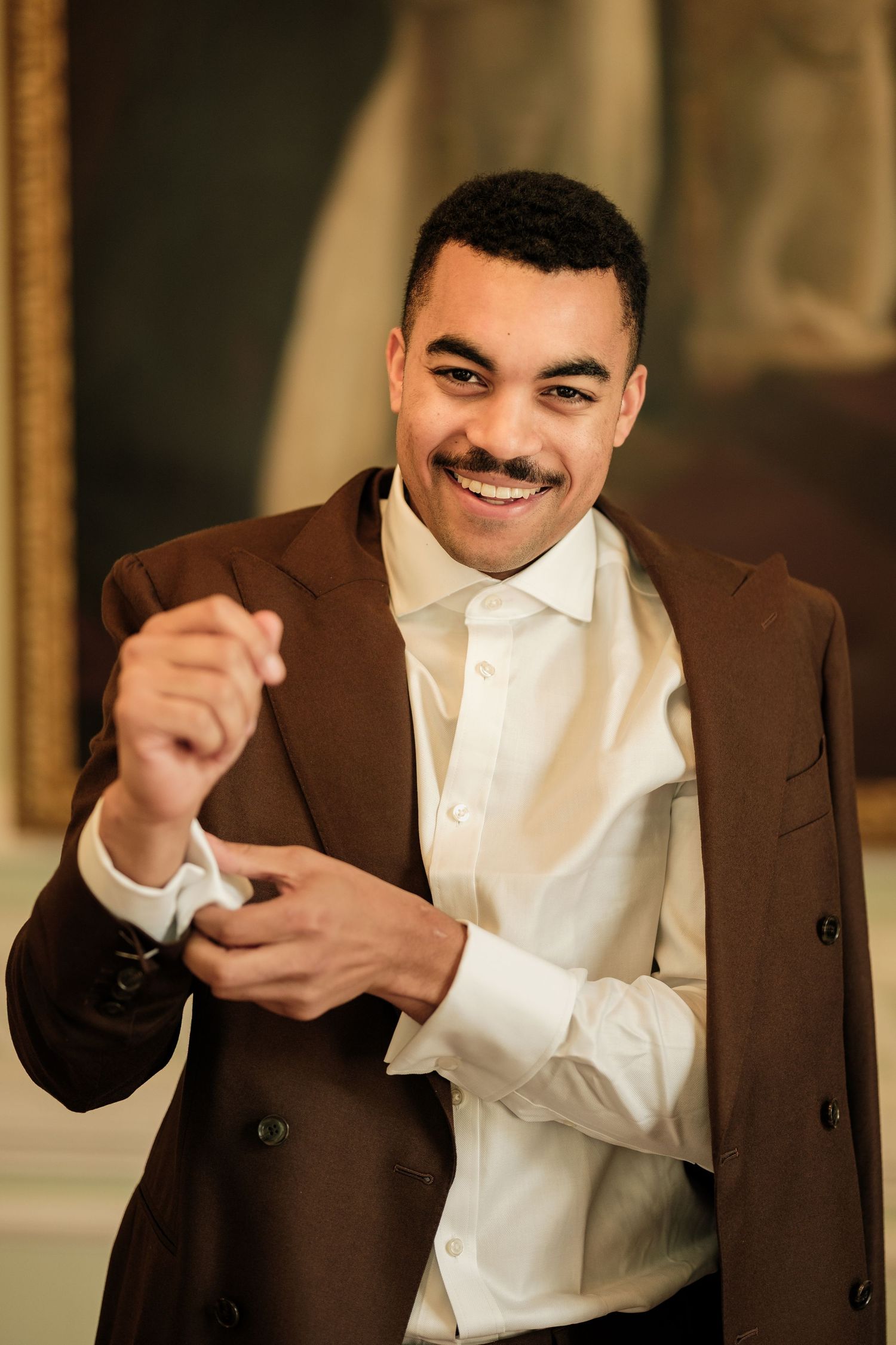 Smiling man in a brown suit jacket and white dress shirt adjusts his cuff while standing in an elegant room with a gilded frame behind him.