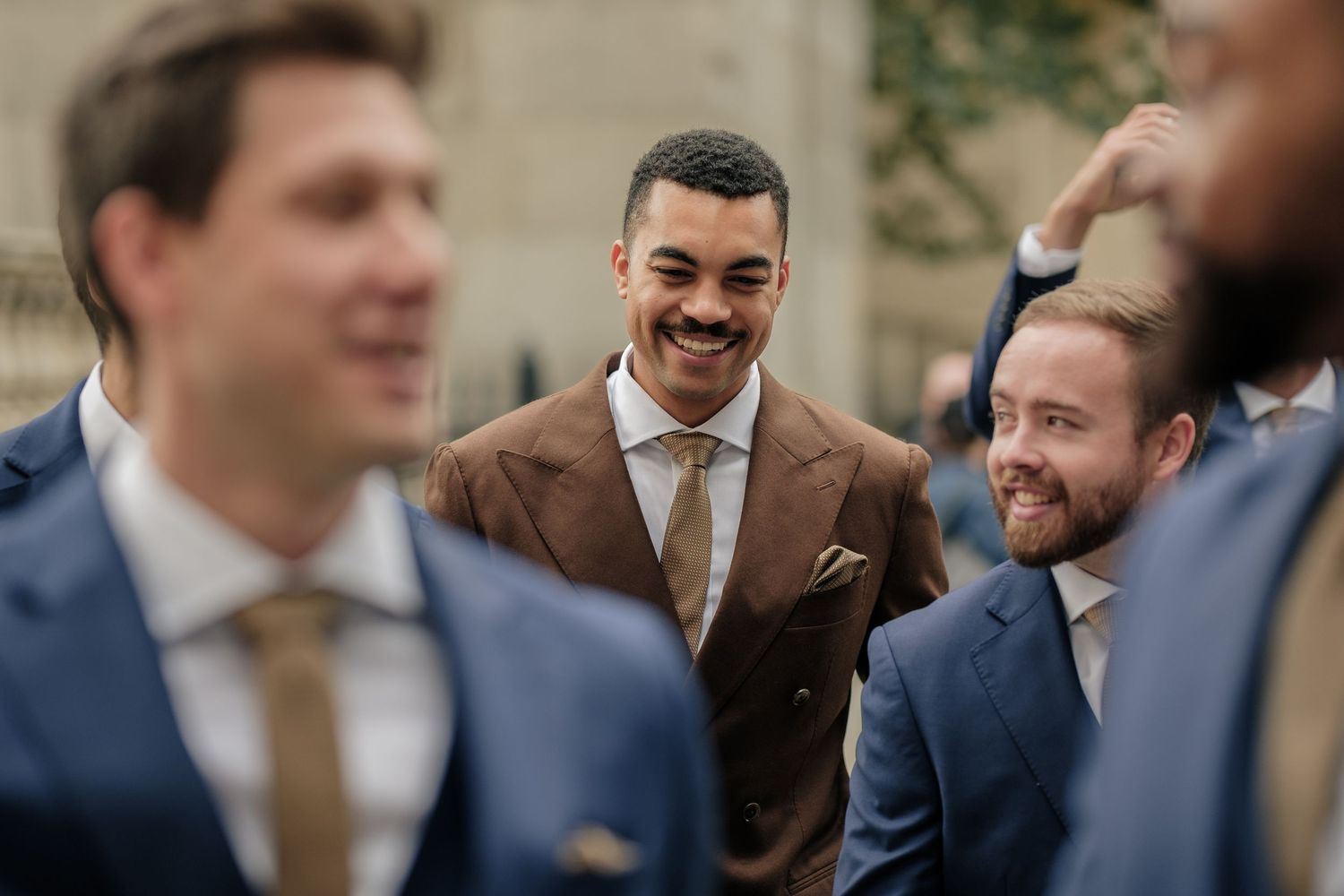 Smiling groomsmen in tailored suits and ties laugh together outdoors, with a man in a brown suit and pocket square at the center.