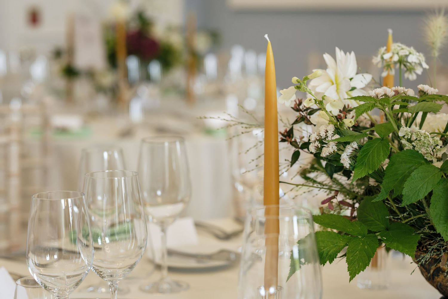 Elegant wedding table setting with tall golden taper candles, white floral arrangement with greenery, and crystal wine glasses.