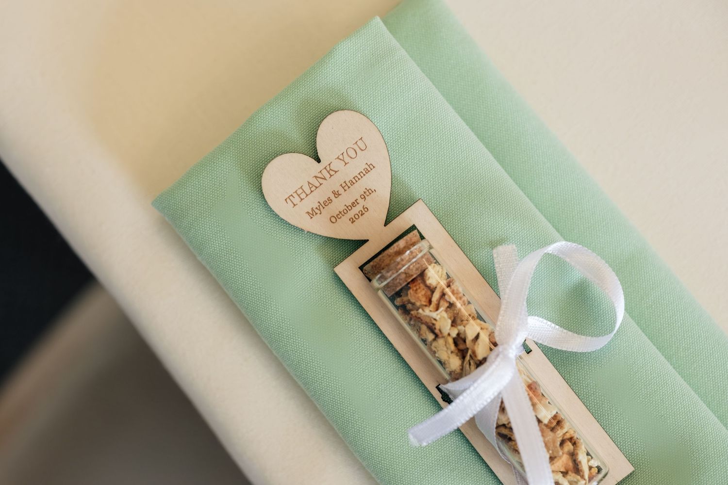Wedding favor with a wooden heart thank you tag and a ribbon-tied gift vial on a mint green napkin at a place setting.