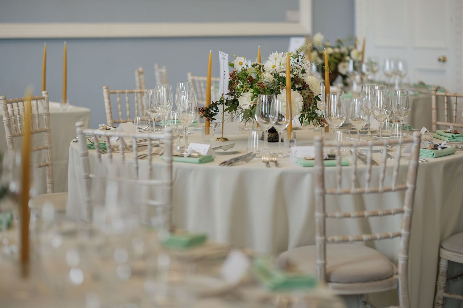 Elegant wedding reception table with white linens, tall candles, floral centerpiece, green napkins, glassware, and white chiavari chairs.