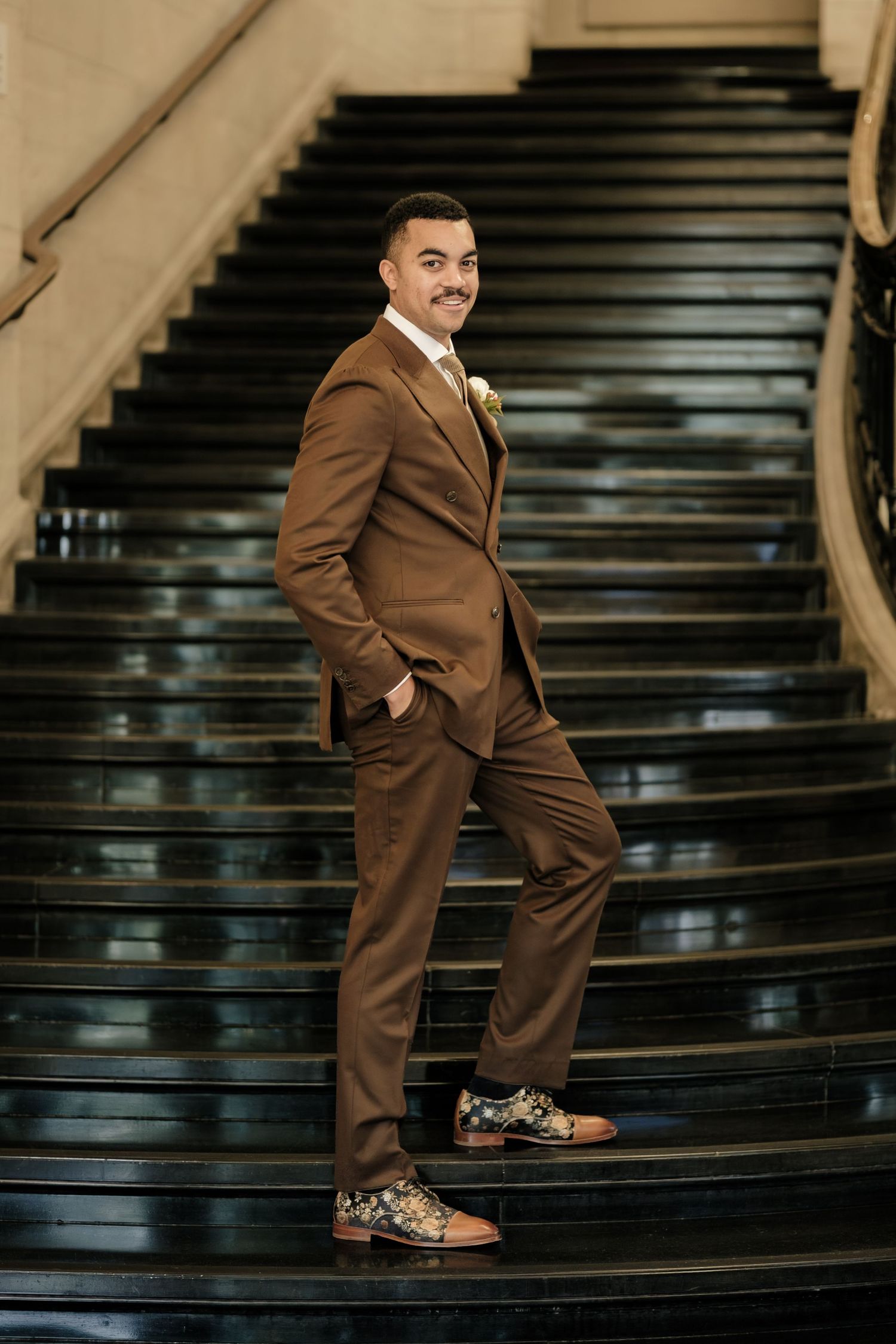 Groom in a brown three-piece suit and patterned floral dress shoes poses with a hand in his pocket on a dark glossy staircase.