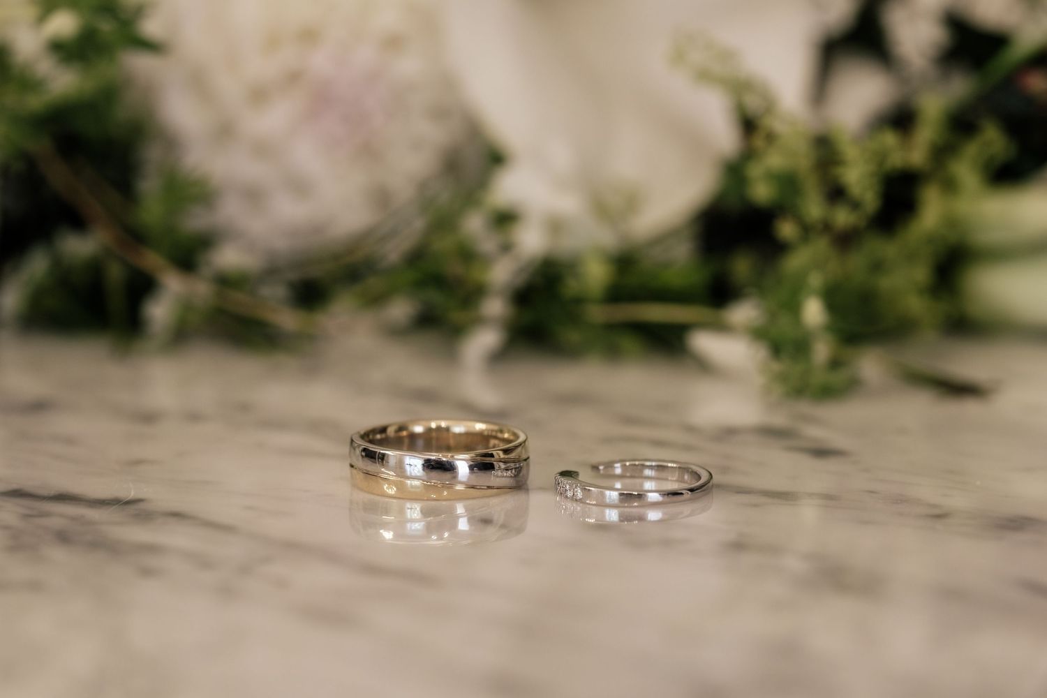 Two wedding bands, one wider with gold detail and one slim silver, rest on a marble surface with soft white flowers and greenery behind.