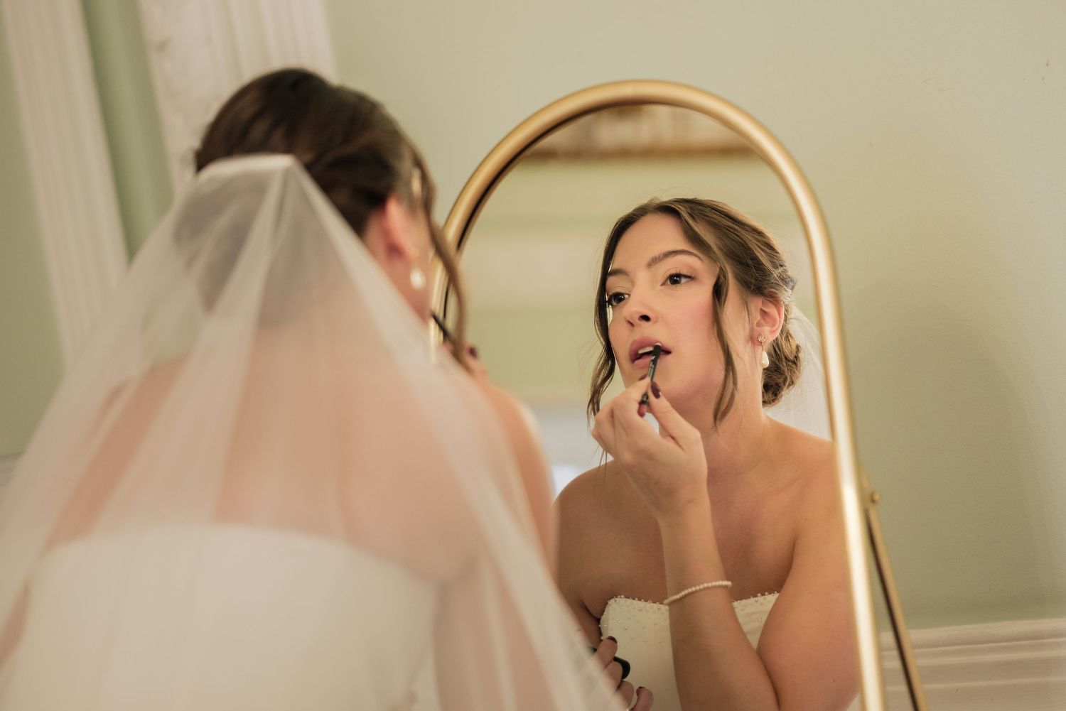 Bride in a white veil and strapless gown applies lipstick while looking into a large gold-framed mirror before her wedding.