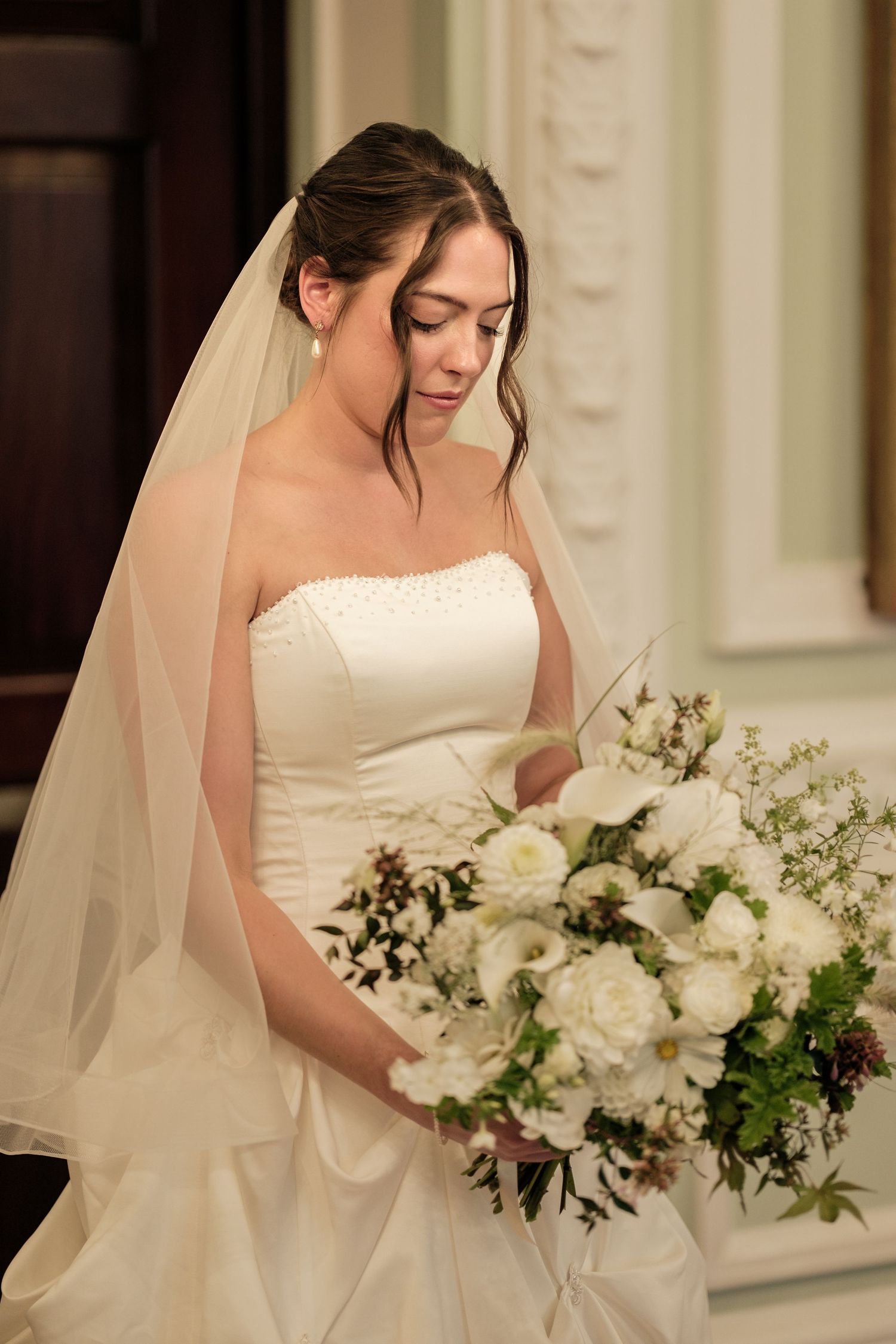 Bride in a strapless white wedding gown and tulle veil looks down at her bouquet of white roses and greenery indoors.