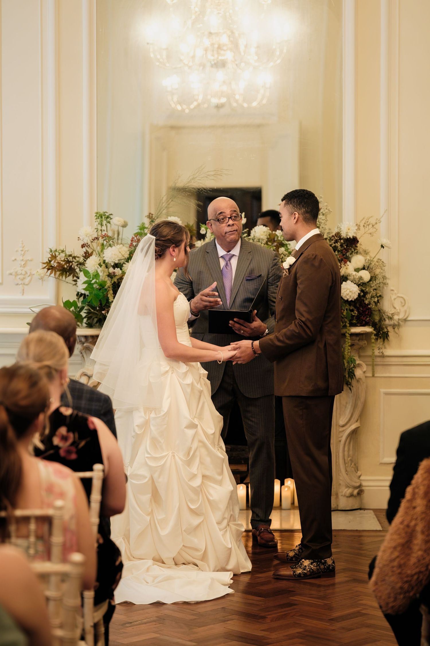Bride in a white ruffled gown and groom in a brown suit hold hands as an officiant leads their wedding ceremony before seated guests.