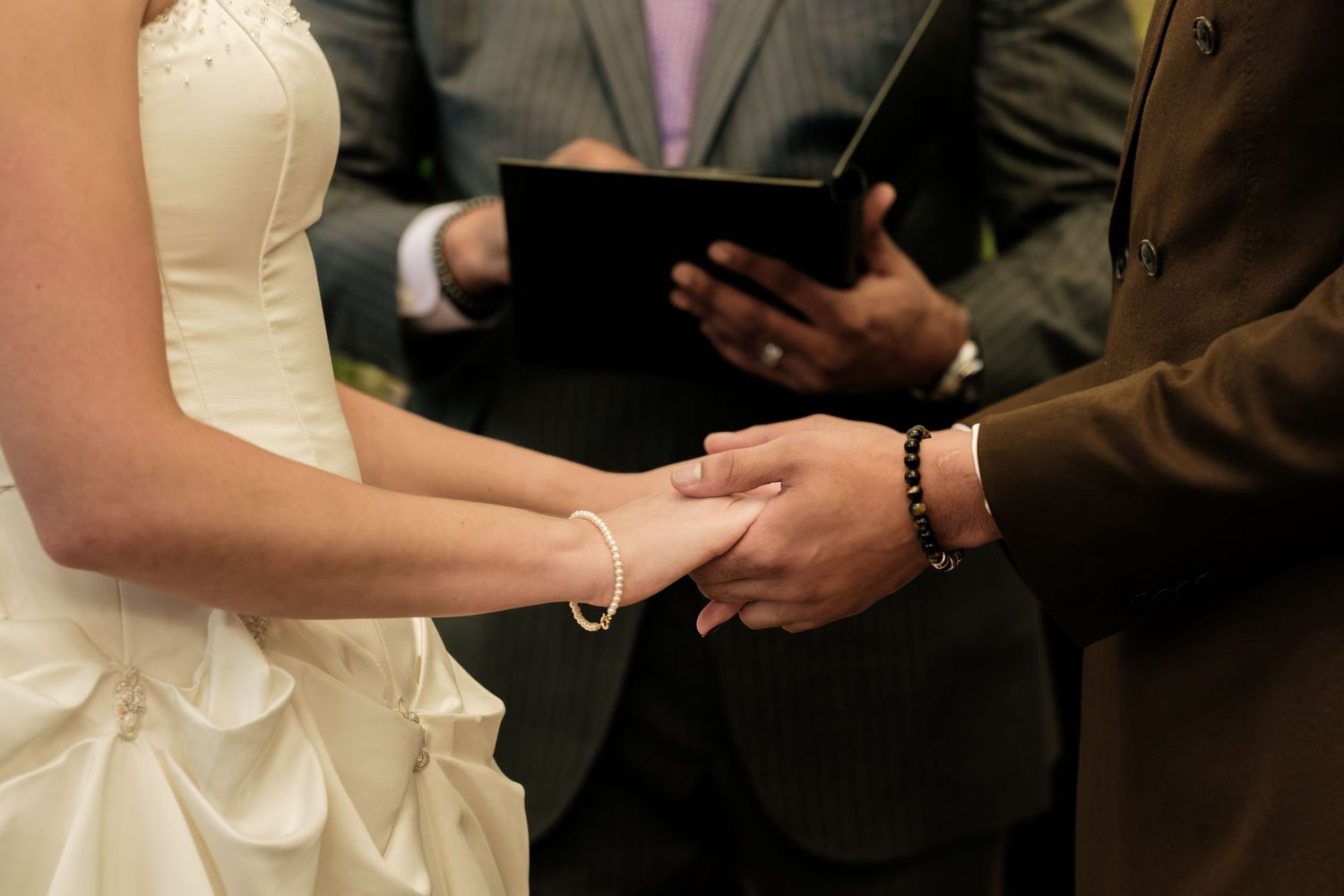 Bride in a white gown and groom in a brown suit hold hands during a wedding ceremony as an officiant reads from a black book.
