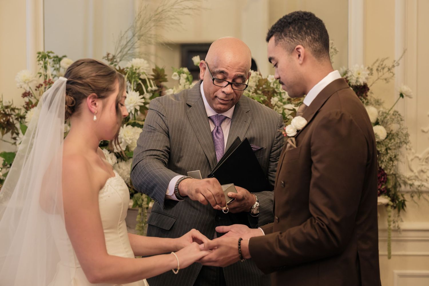 A bride in a white gown and a groom in a brown suit hold hands while an officiant in glasses leads their wedding ceremony among flowers.