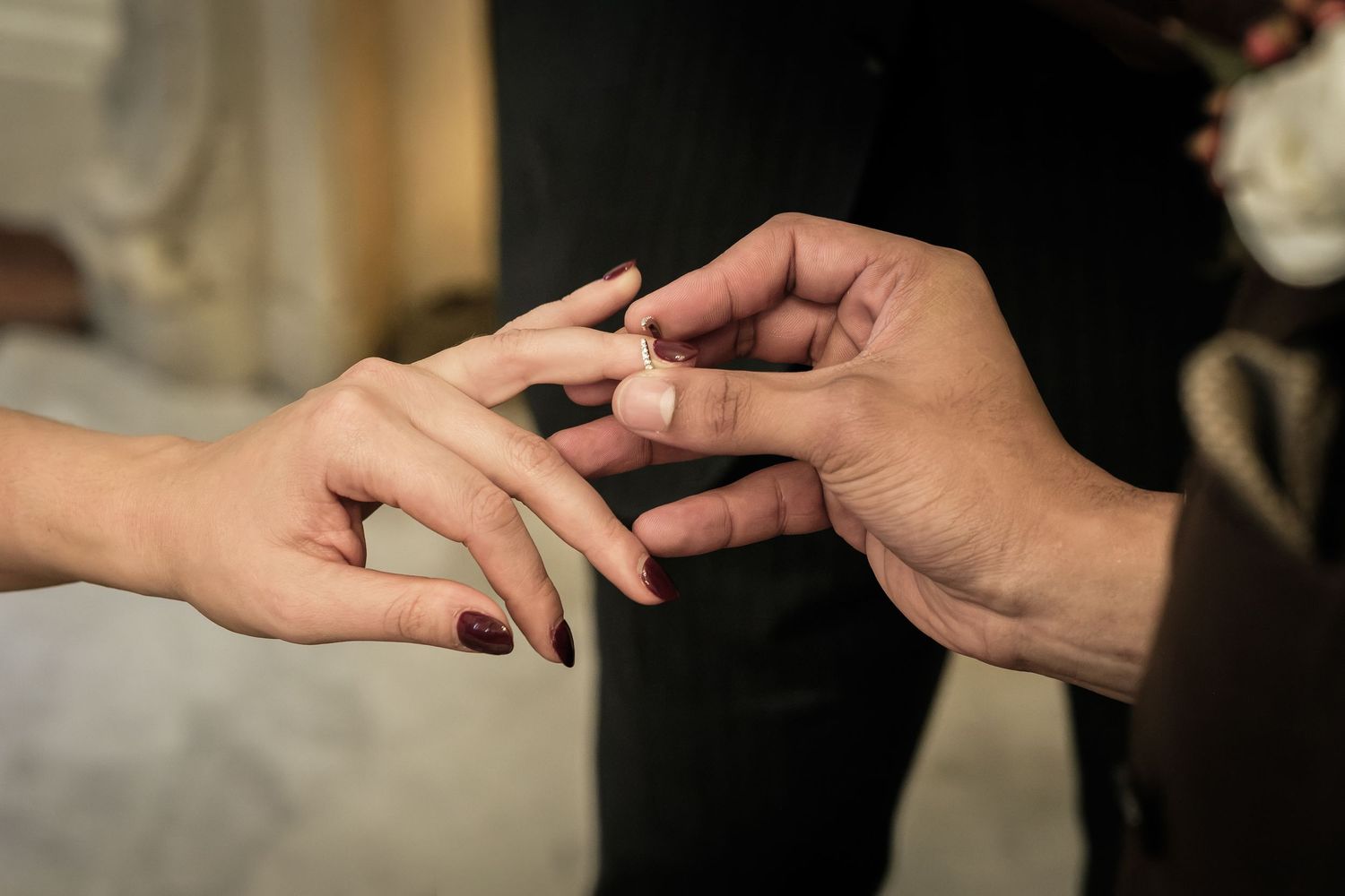 A man's hand slides a diamond engagement ring onto a woman's finger, her nails painted dark red, during a romantic proposal.