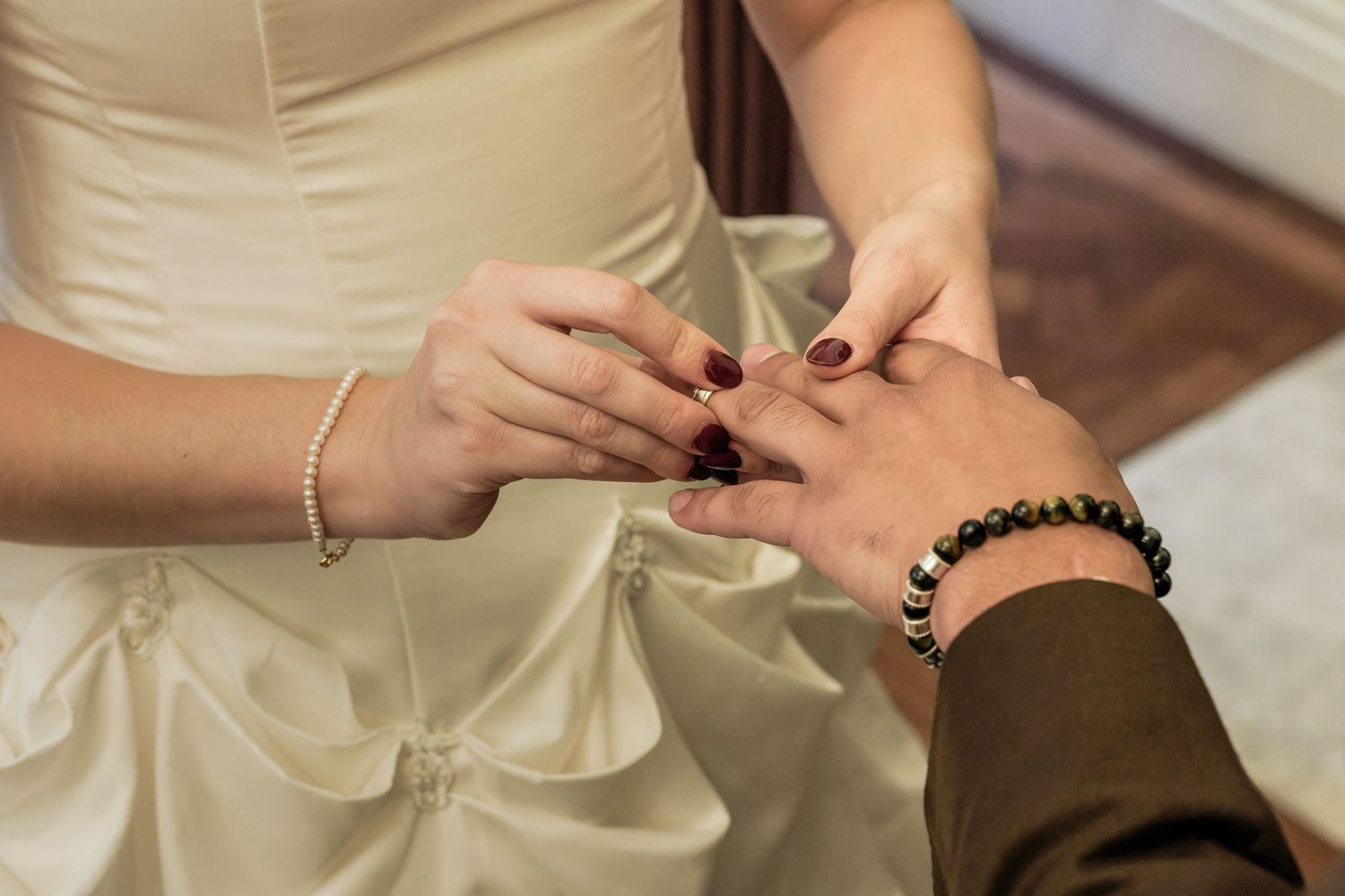 Bride in a white satin gown slips a wedding ring onto the groom's finger during a wedding ceremony exchange of vows.