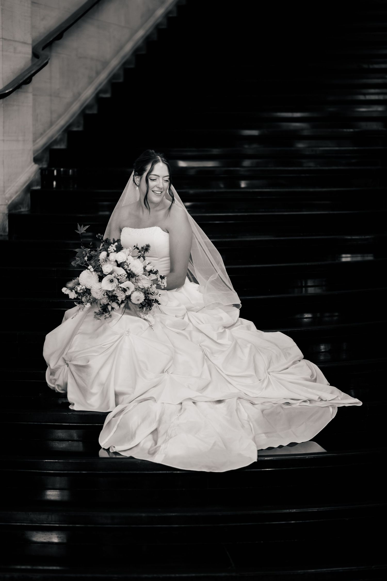 Black and white photo of a smiling bride in a flowing gown and veil sitting on dark stairs holding a bouquet of flowers.