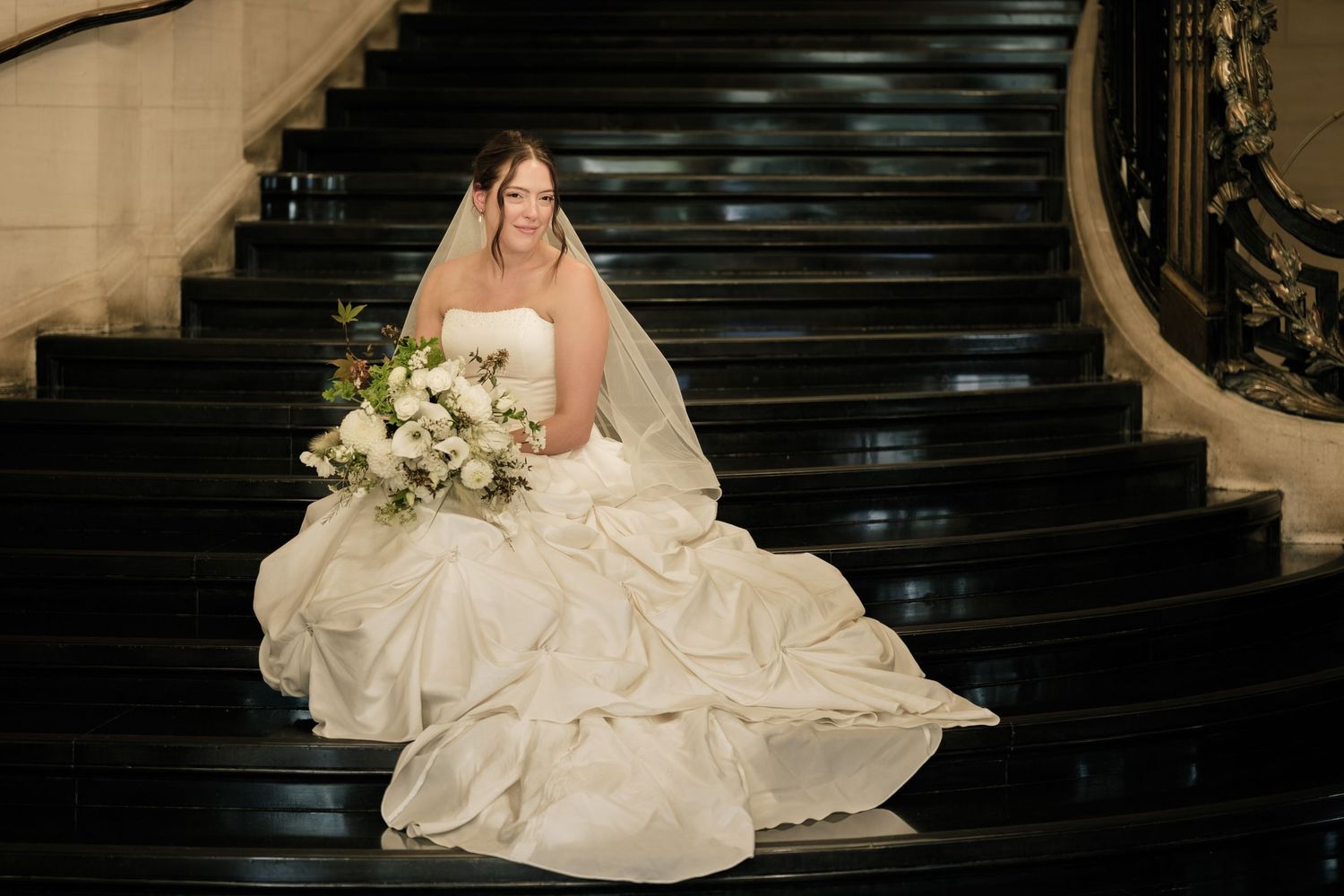 Bride in a white strapless wedding gown and veil sits on a dark marble staircase holding a white floral bouquet.