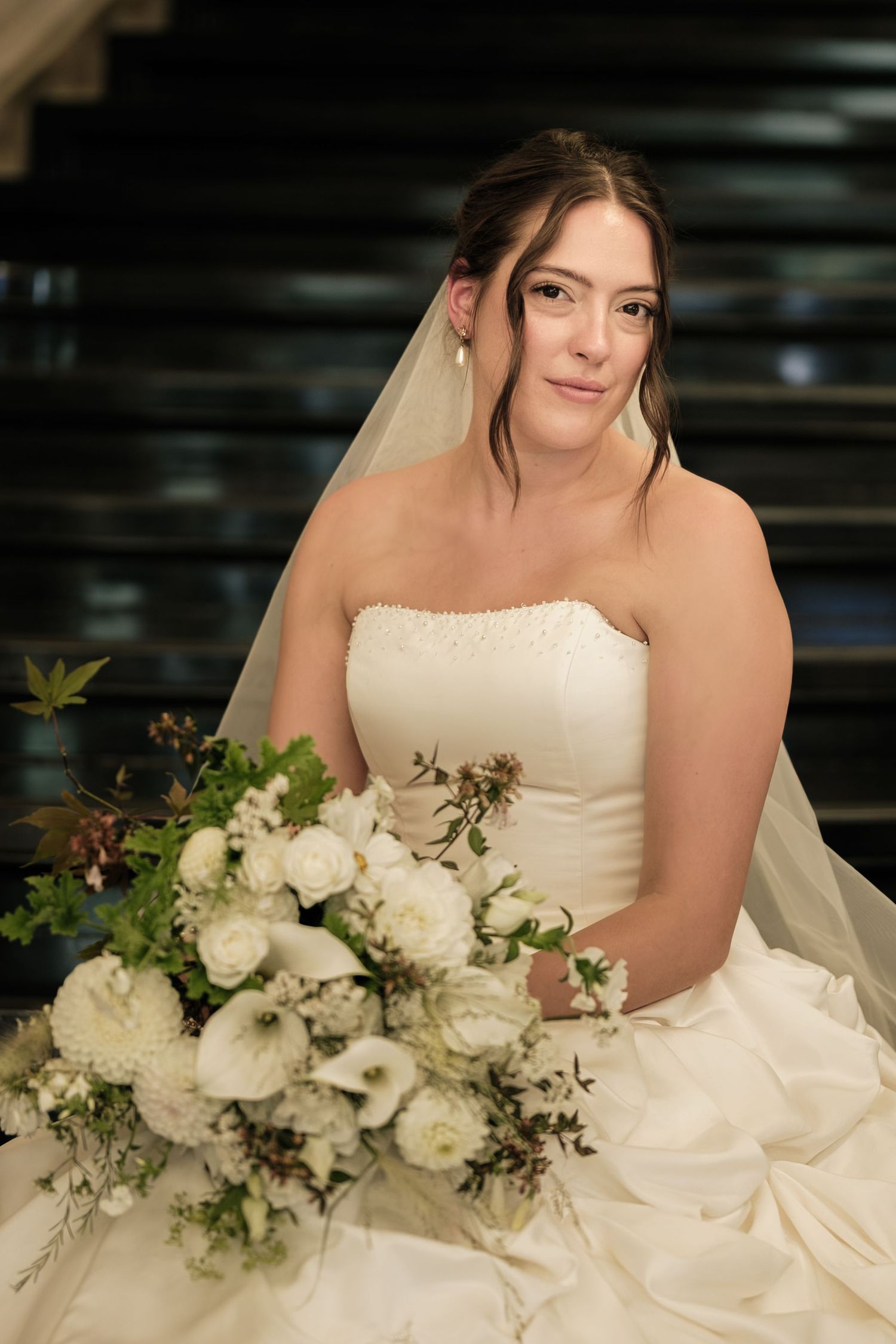 Smiling bride in a strapless white gown and veil holds a lush bouquet of white flowers and greenery while seated on a dark staircase.