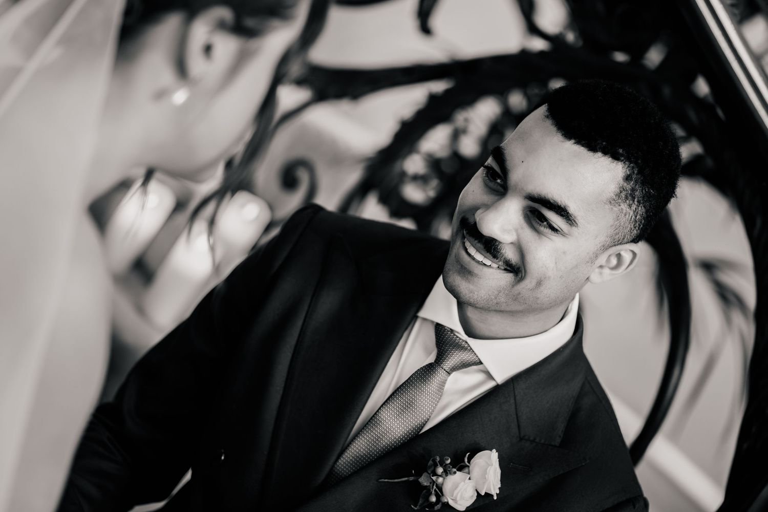 Smiling groom in a dark suit with a floral boutonniere gazes lovingly at his bride in a black and white wedding portrait.