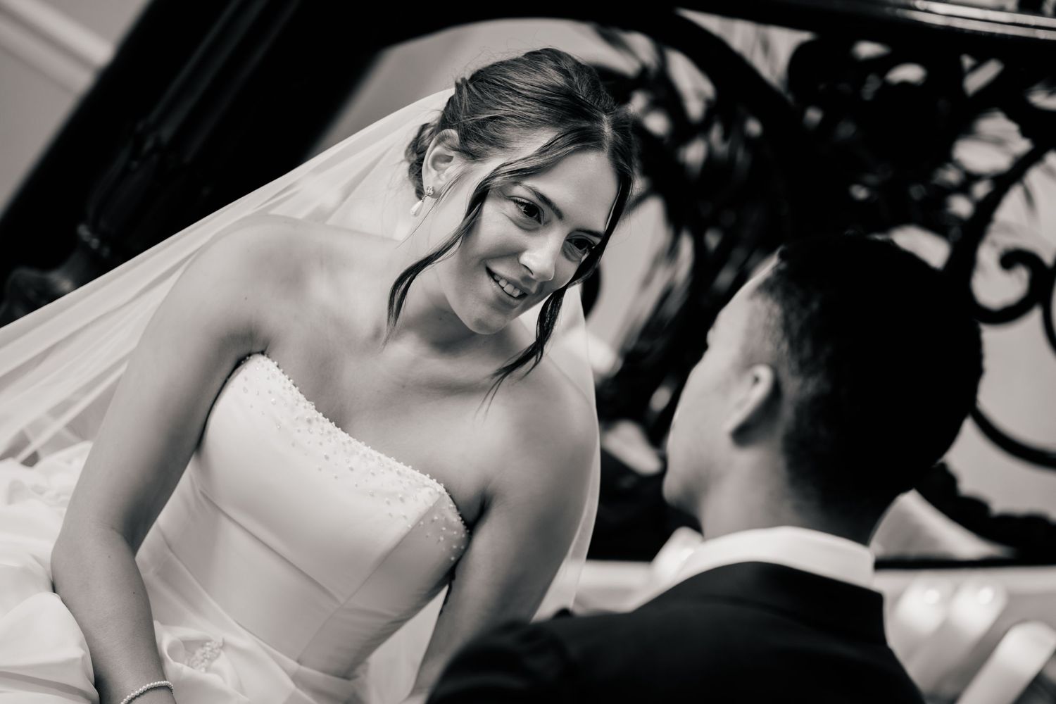 Black and white photo of a smiling bride in a strapless gown and veil looking warmly at her groom in a dark suit during a wedding ceremony.