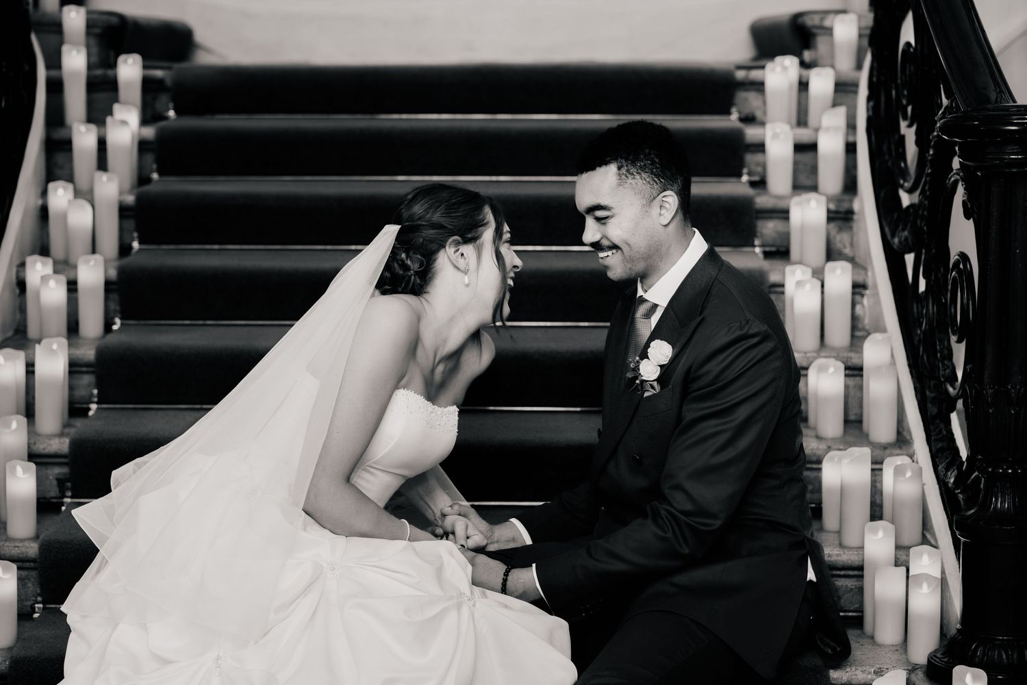 Black and white photo of a bride and groom sitting close on a candlelit staircase, smiling at each other on their wedding day.