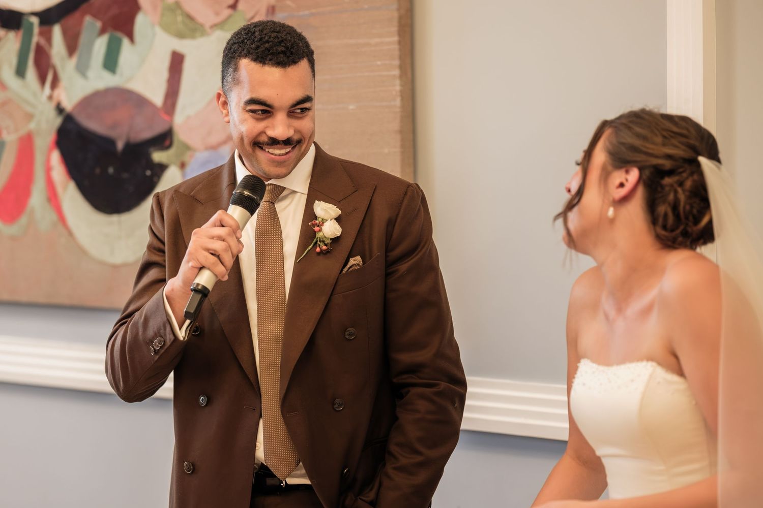 Smiling groom in a brown suit with a floral boutonniere speaks into a microphone as his bride in a white strapless dress looks on.