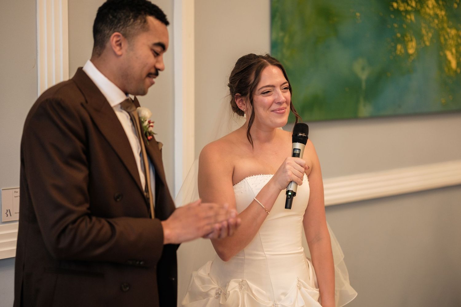 Smiling bride in a white strapless gown holds a microphone while a groom in a brown suit speaks beside her during a wedding reception toast.