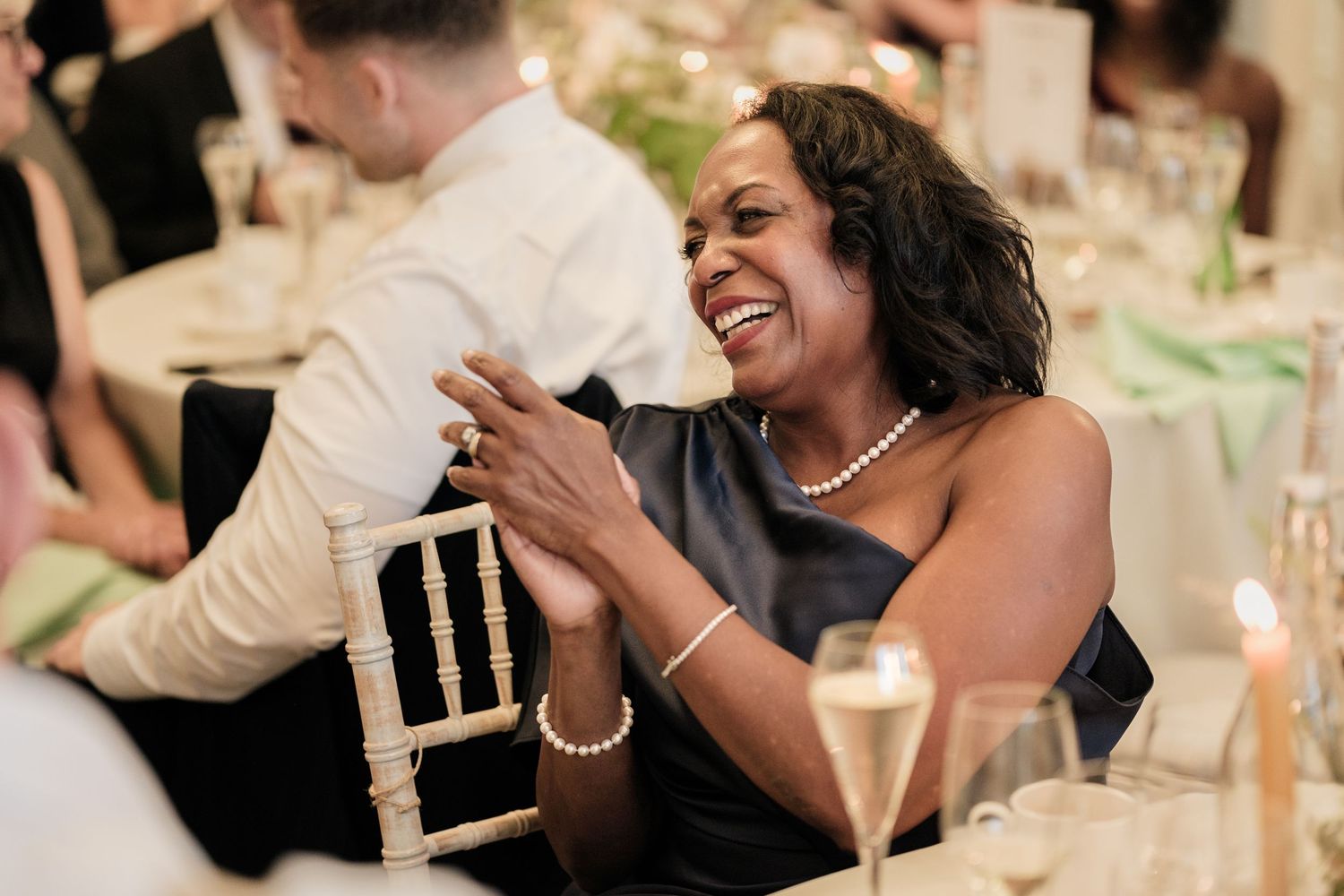 Smiling woman in a black dress and pearl jewelry laughs at a elegant wedding reception table with champagne glasses and candles.
