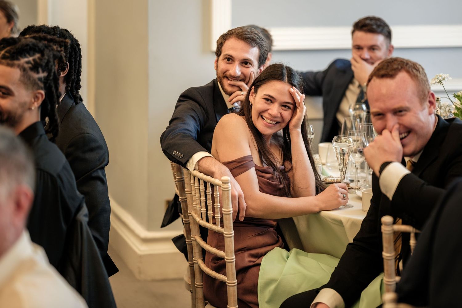 Smiling wedding guests in formal attire laugh together at a decorated dining table during a reception.