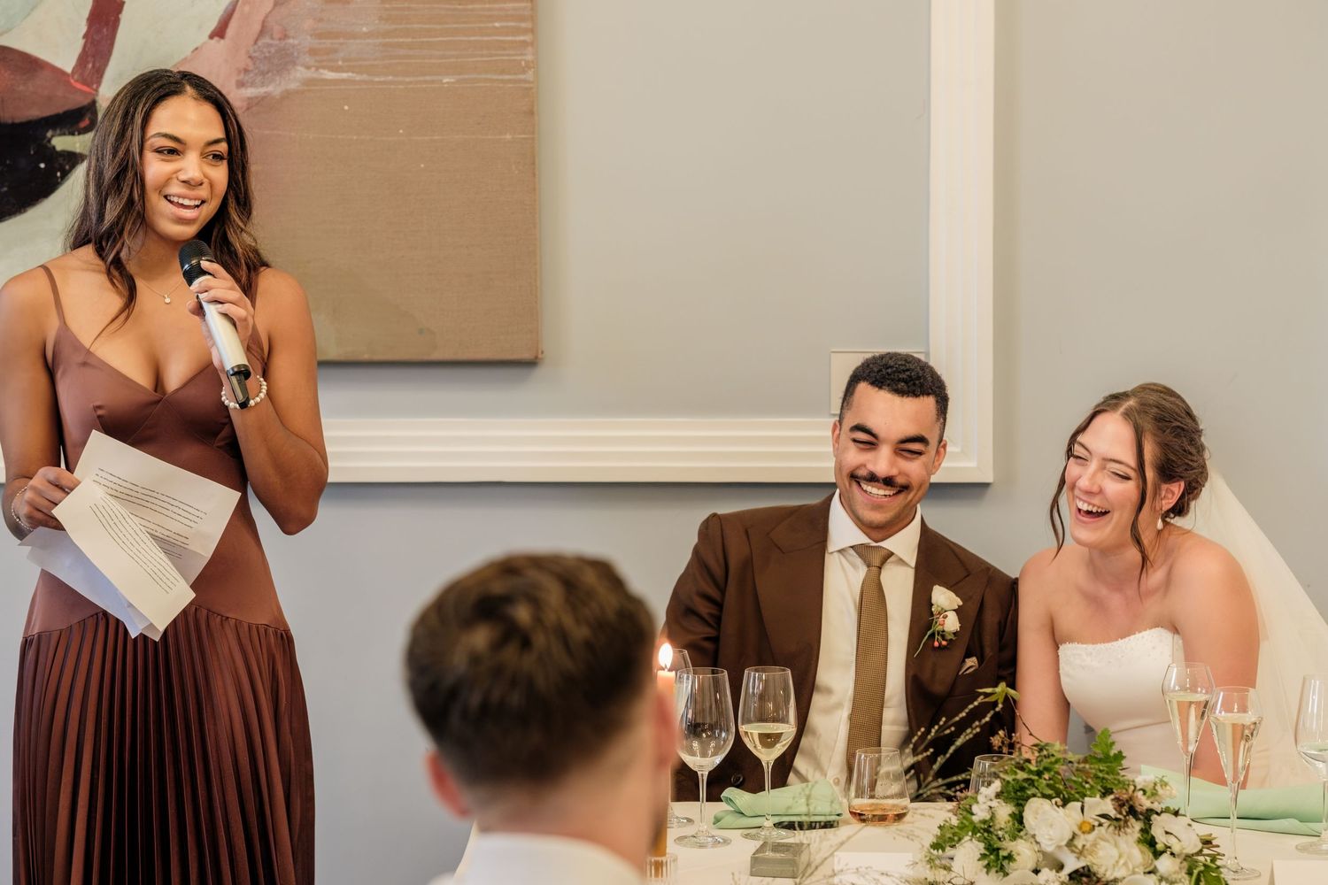 Bridesmaid in a pleated bronze dress gives a wedding toast with a microphone while the smiling bride and groom laugh at the reception table.