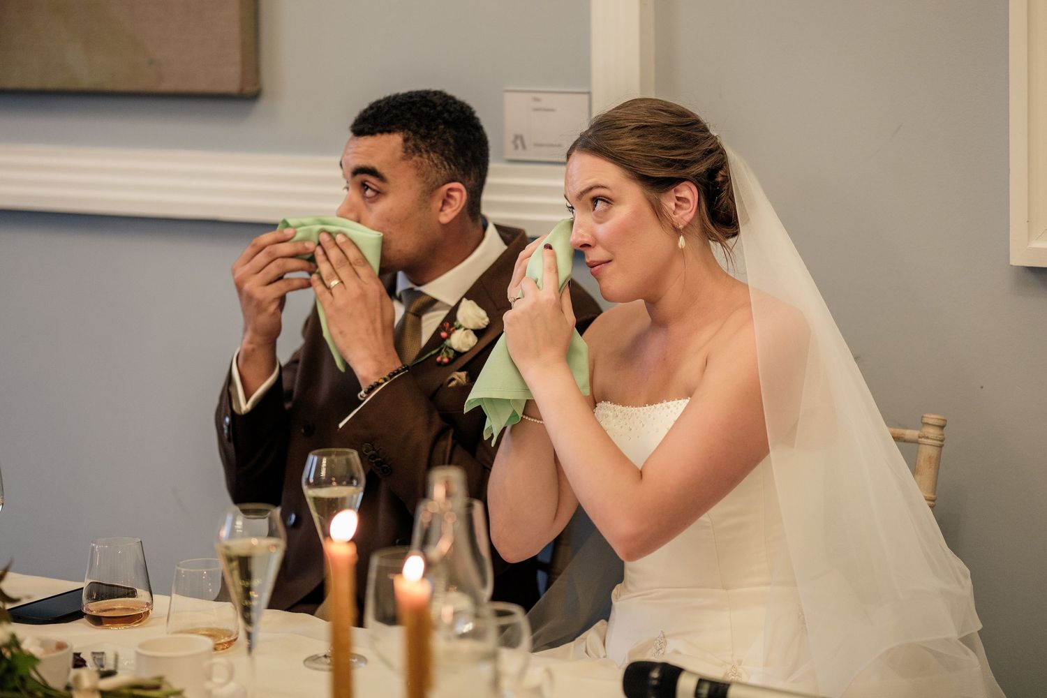Emotional bride in a white gown and veil and groom in a suit dab their tears with green napkins at a candlelit wedding reception table.