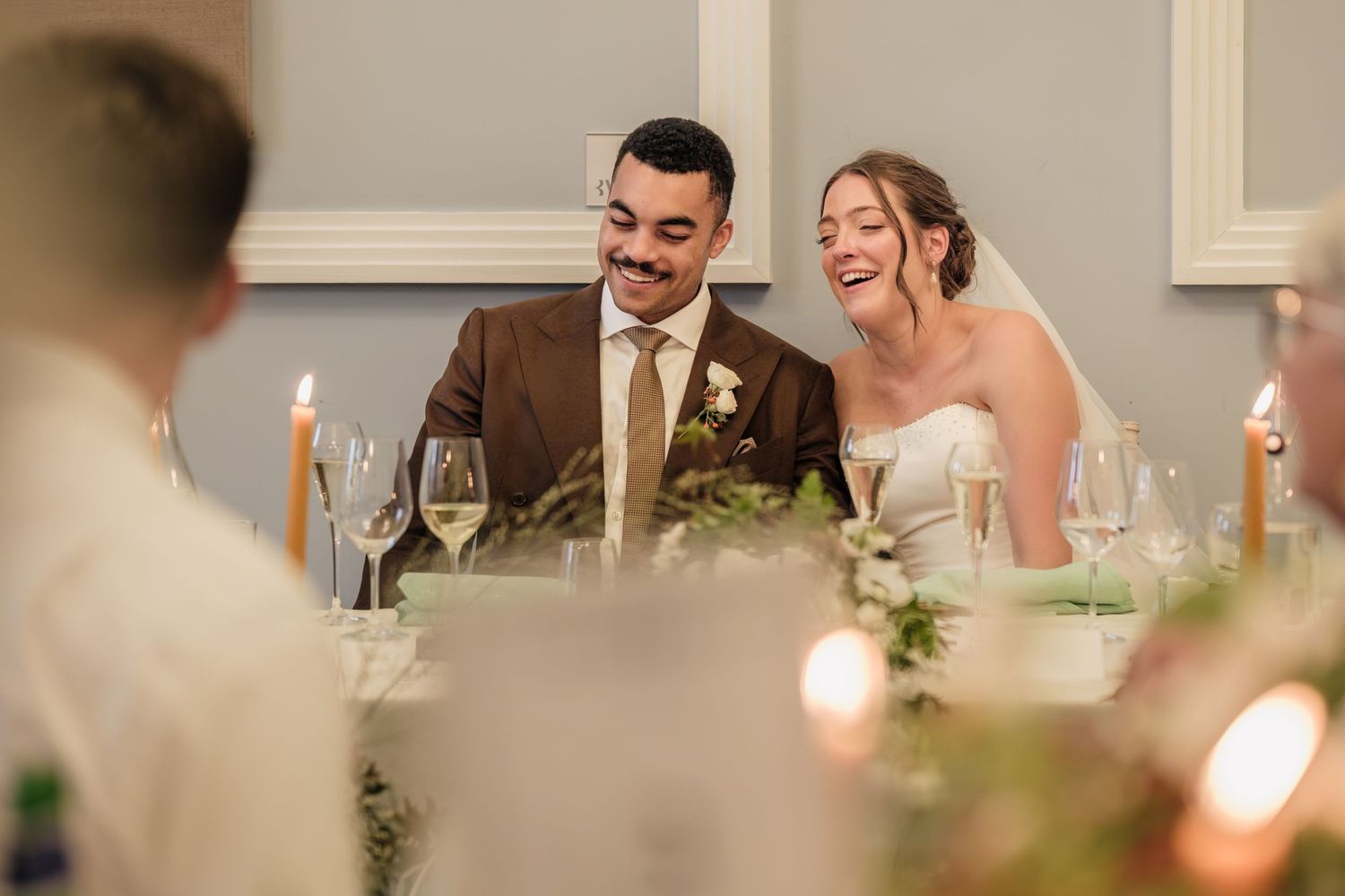 Smiling bride and groom in a brown suit sit side by side at a candlelit wedding reception table with champagne glasses and greenery.