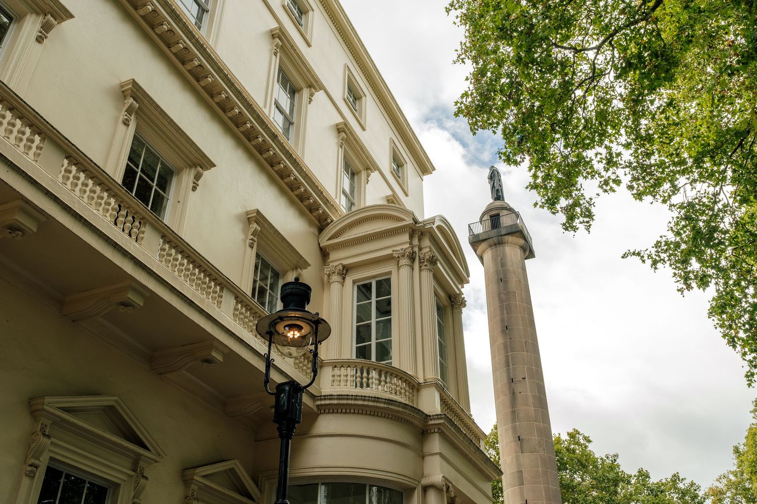 Cream-colored neoclassical building with balustraded balcony and ornate street lamp beside a tall stone monument column and green trees.