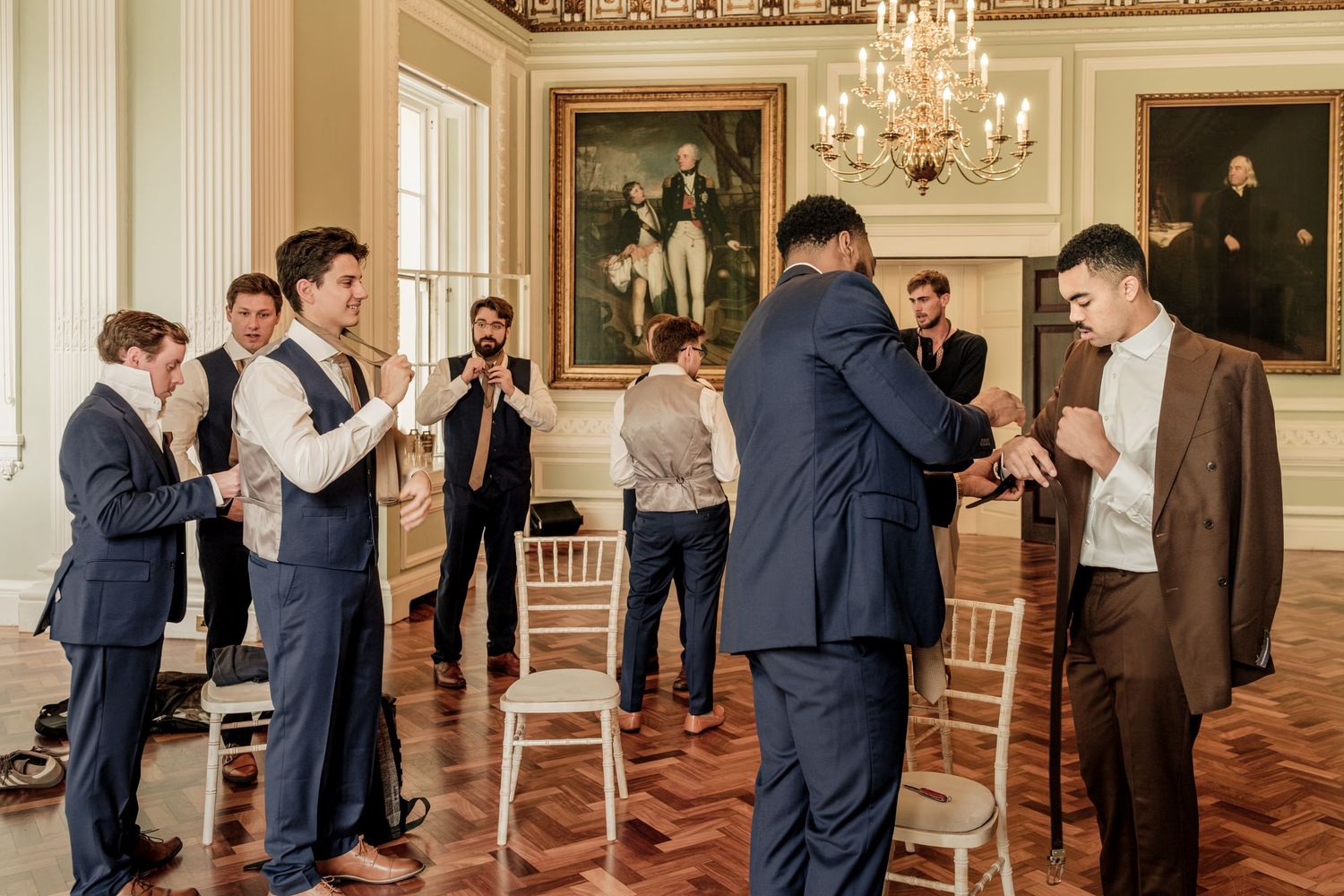 Groomsmen in suits chat and adjust their attire in an elegant ballroom with parquet flooring, chandeliers, and framed portraits.
