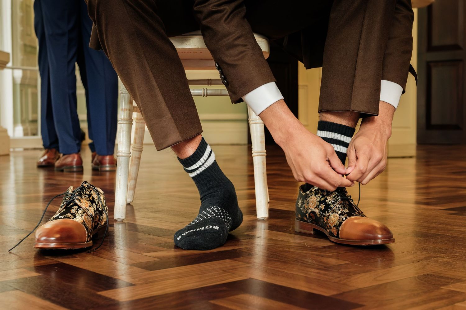 Man in brown trousers seated on a chair tying a floral brogue shoe, wearing striped black socks on a polished parquet wood floor.