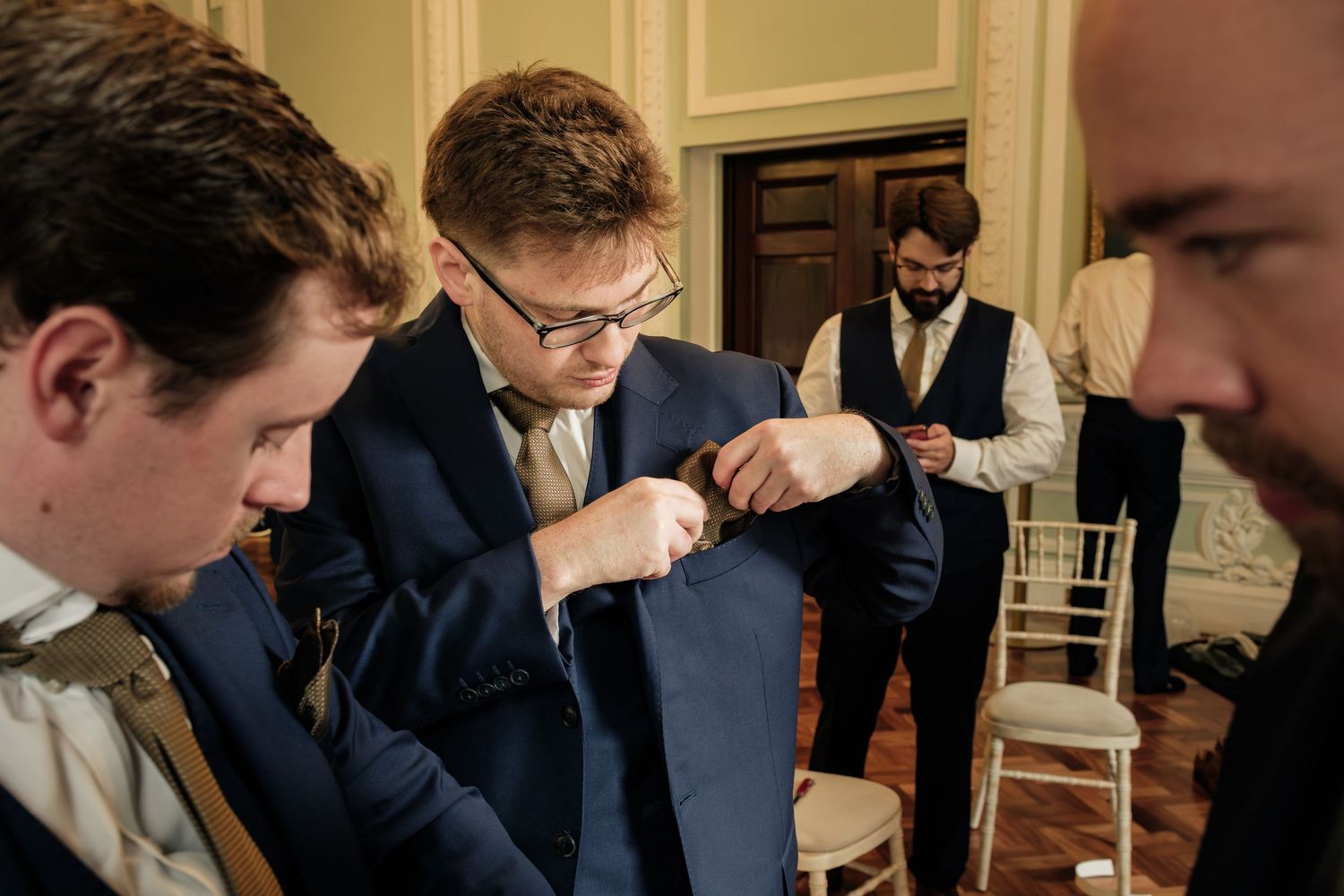 Groomsman in glasses and a navy suit pins a boutonniere to his lapel while other men in formal wear stand in an elegant room.
