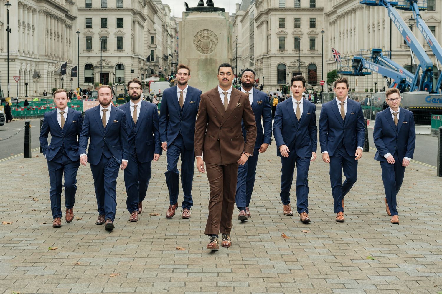 Group of men in tailored navy suits walking in formation behind a leader in a brown suit through a sunlit city square.