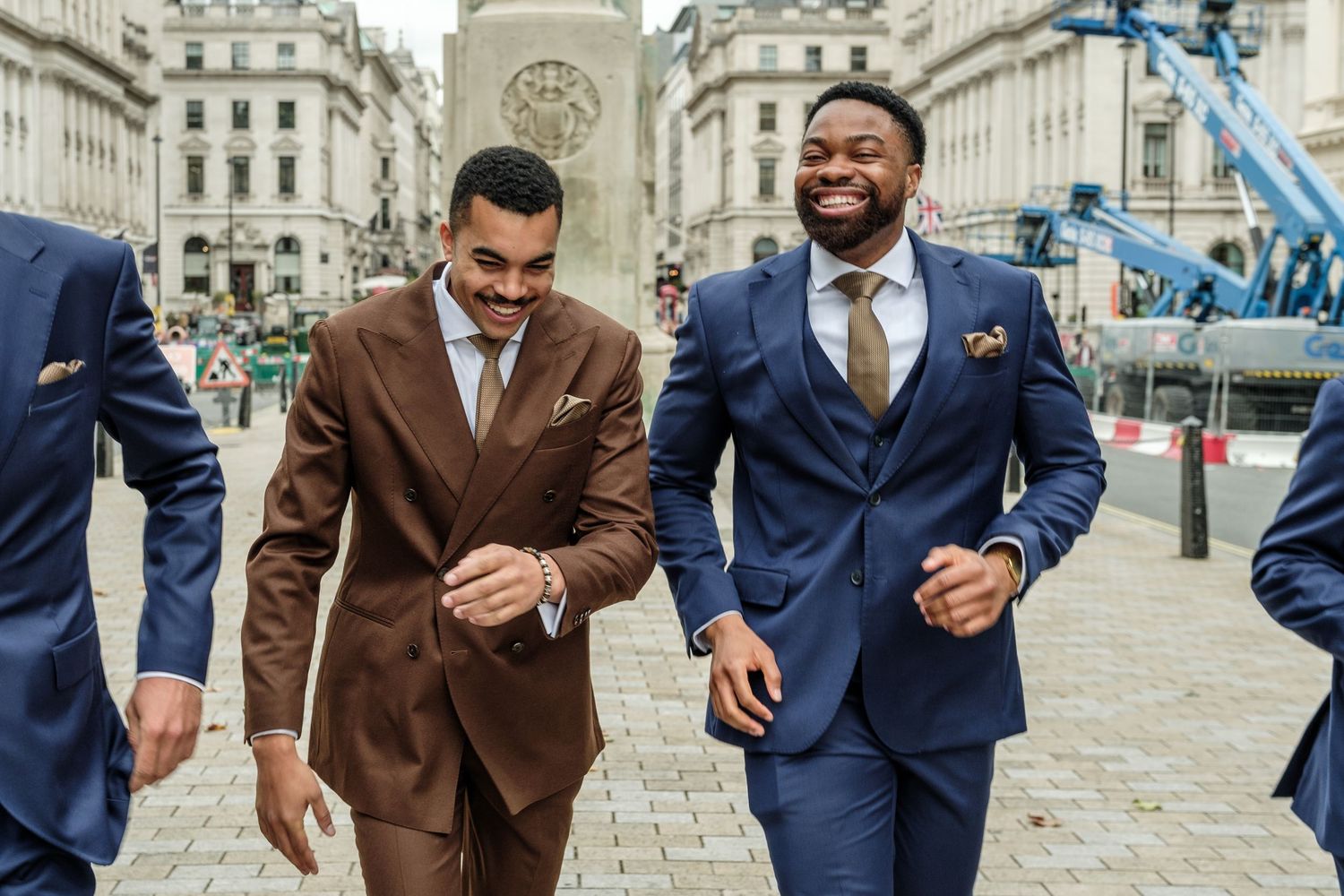 Three smiling men in tailored brown and navy suits walk together along a sunlit cobblestone city street lined with historic buildings.
