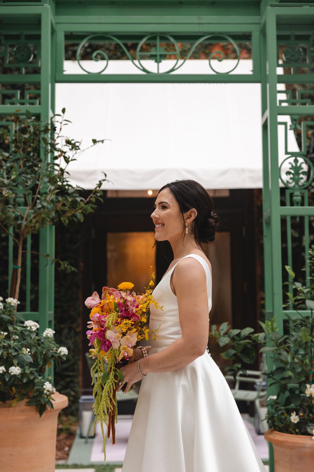 Smiling bride in a sleeveless white gown holds a colorful bouquet while standing beneath a green garden trellis with potted white flowers.