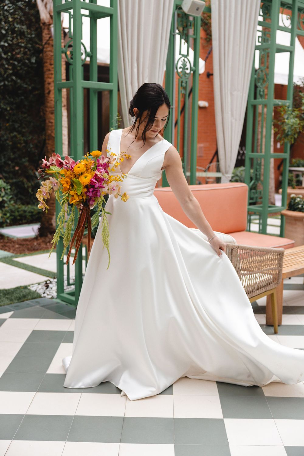 Bride in a white satin V-neck gown holds a colorful orange and pink bouquet while standing on a green and white checkered patio.