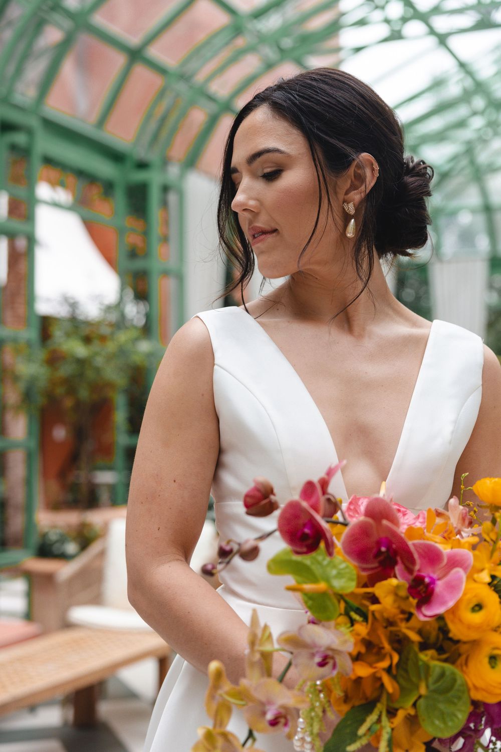 Bride in a white V-neck gown holds a colorful bouquet of pink orchids and orange blooms inside a bright greenhouse venue.
