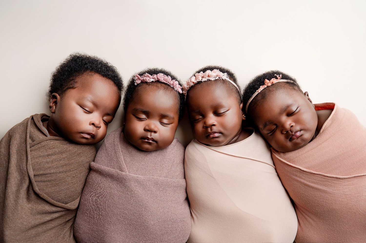 Four sleeping newborn babies swaddled in pink and mauve wraps, wearing floral headbands, posed together in a row.