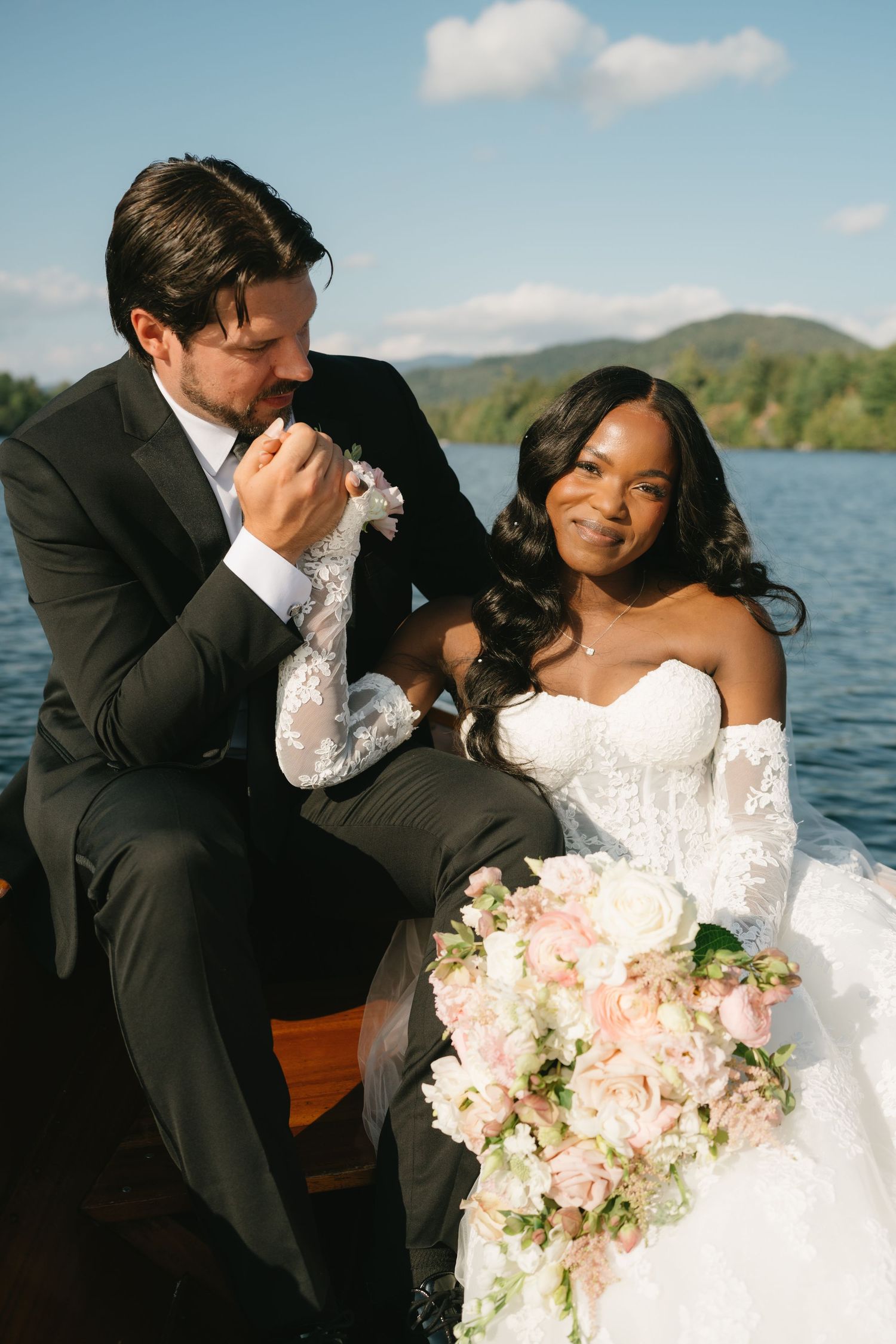 Interracial couple in wedding attire sit on a boat, groom kissing bride's hand, bride holding pink bouquet.