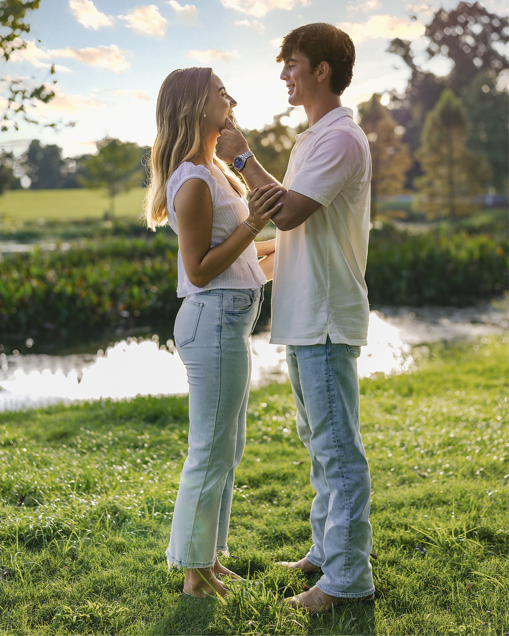 Couple dancing together on green grass near a pond, wearing white tops and light blue jeans at sunset.