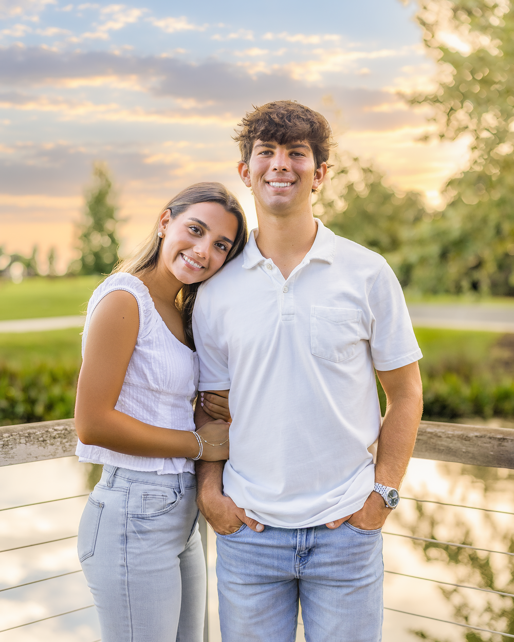 Two young adults in white tops and jeans smile together outdoors at sunset near a wooden fence.
