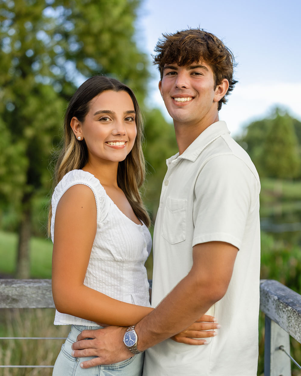 A young couple smiles and poses together outdoors near a fence with green trees in the background.