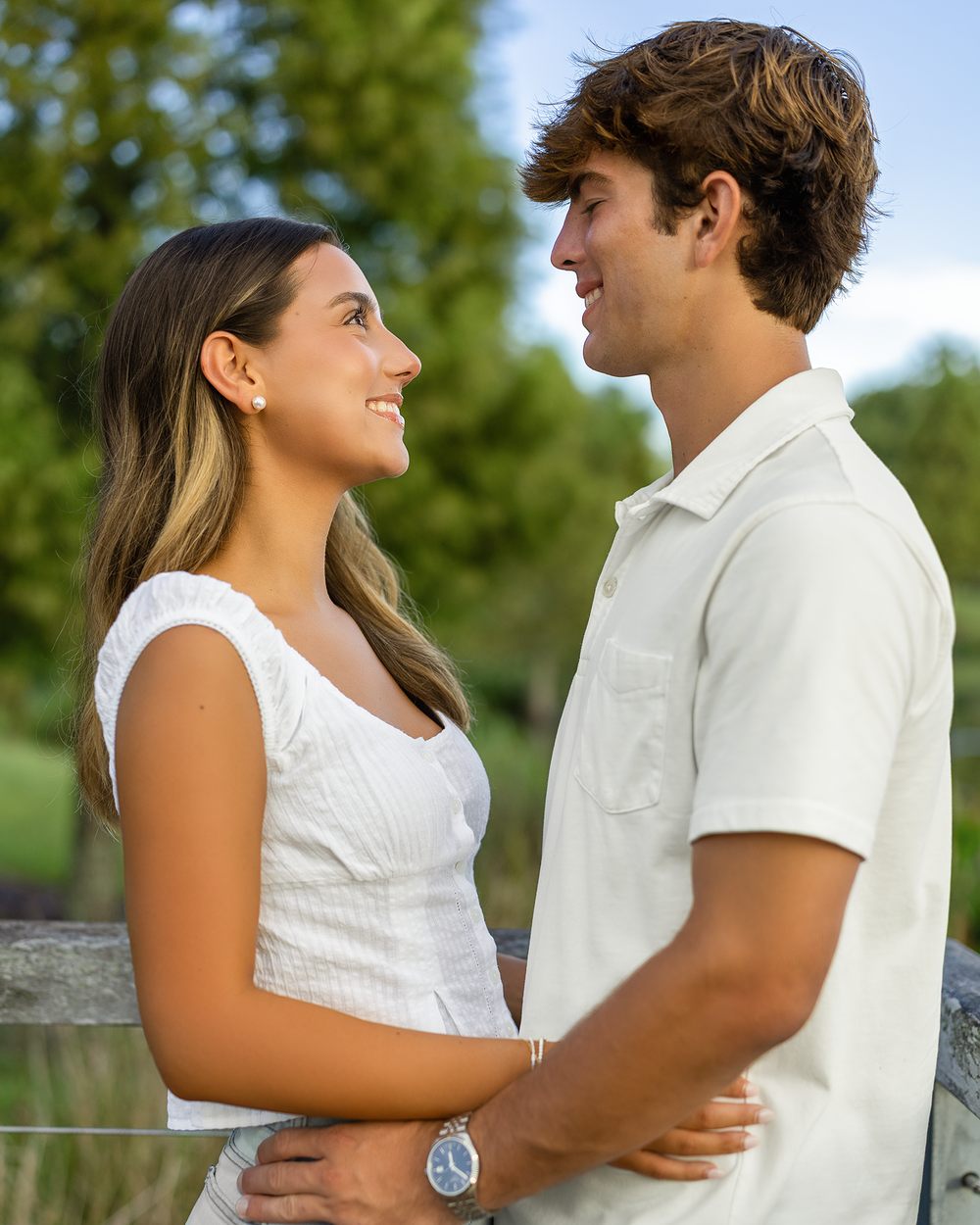 A young couple in white outfits smiling at each other outdoors with green trees in the background.