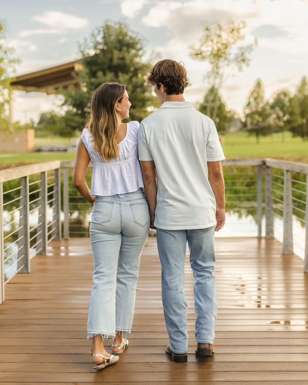 Couple walking hand-in-hand on a wooden bridge, wearing matching white tops and light blue jeans outdoors.