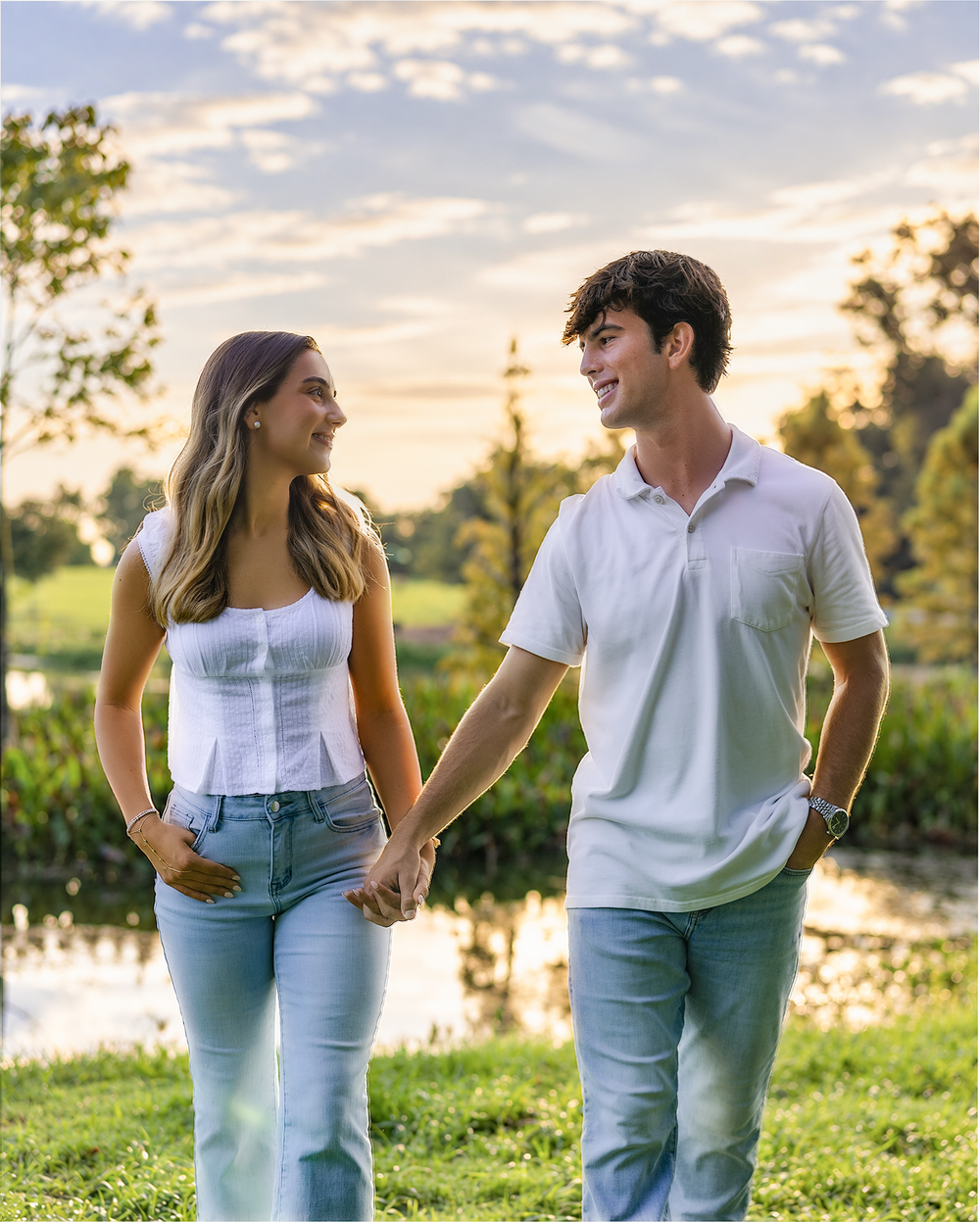 A young couple holds hands and smiles at each other while walking in a lush green park at sunset.