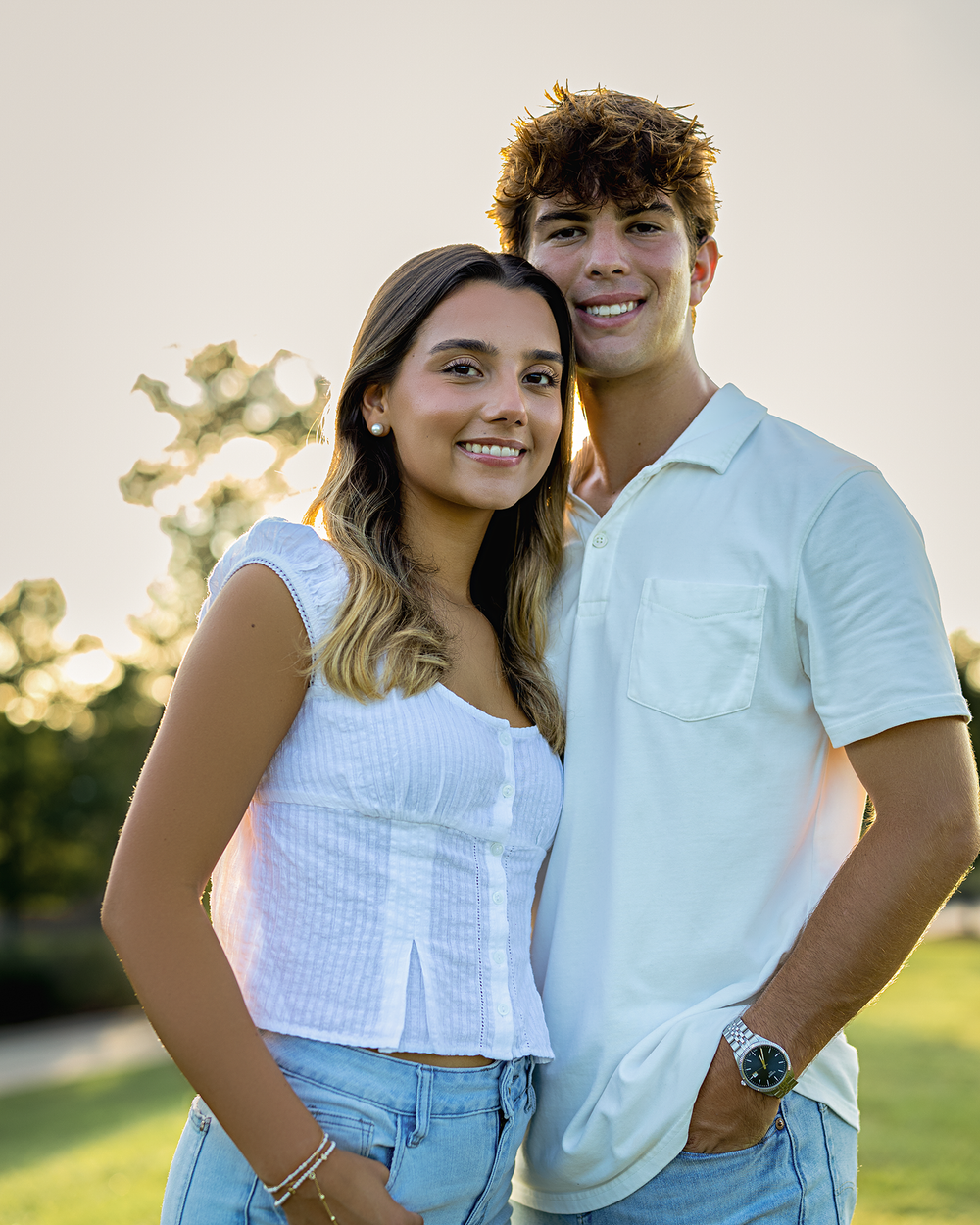 A young couple smiling outdoors at golden hour, both wearing white tops and jeans in a sunny park setting.