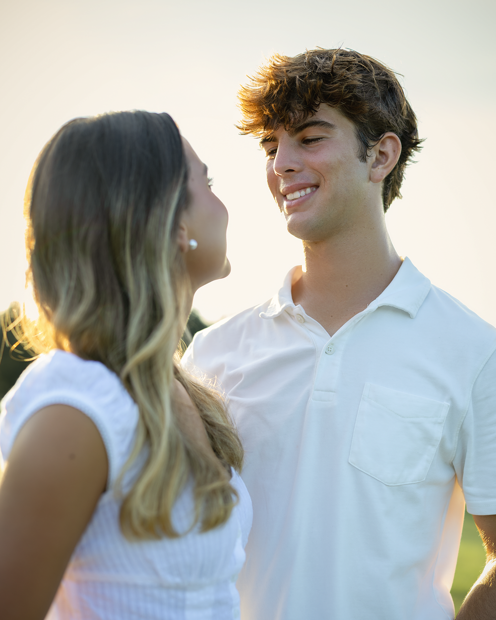 A smiling young couple in white outfits share a tender moment outdoors during golden hour.