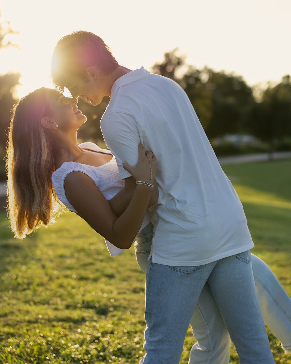 Couple sharing a romantic dip kiss at golden hour in a lush green park, bathed in warm sunset light.