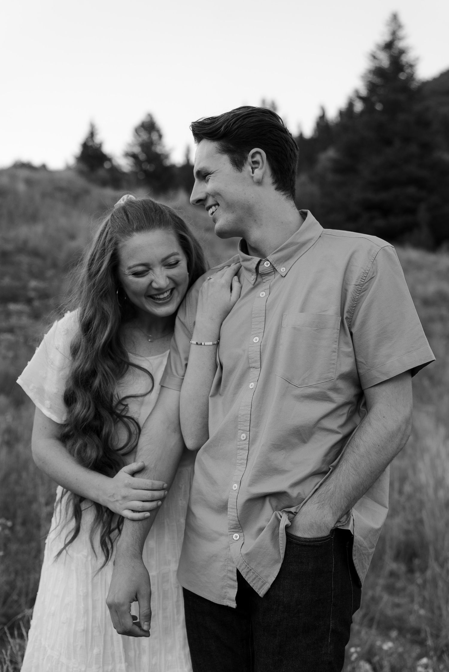 Laughing couple in spring engagement picture outfits. Woman in flutter sleeve white chiffon dress holding man in loose collar button-up